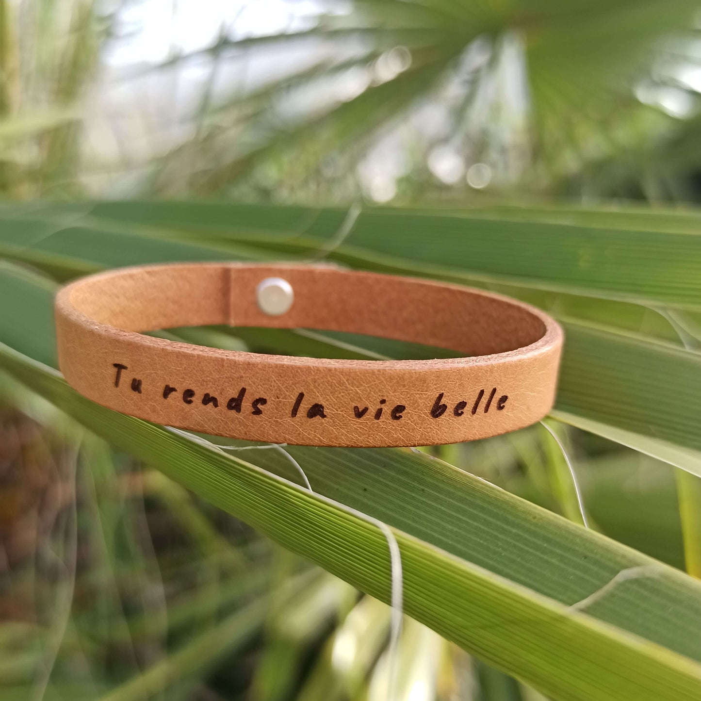 A brown leather bracelet with the text 'Tu rends la vie belle' engraved on it.