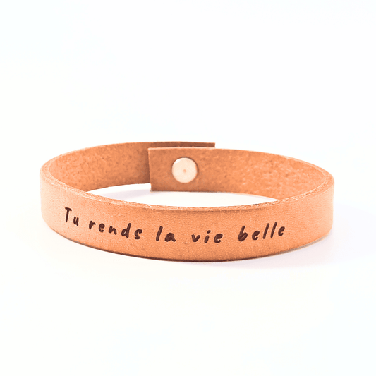 A brown leather bracelet with the text 'Tu rends la vie belle' engraved on it.
