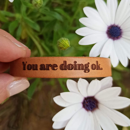 A brown leather bracelet with the text 'You are doing ok.' engraved on it.