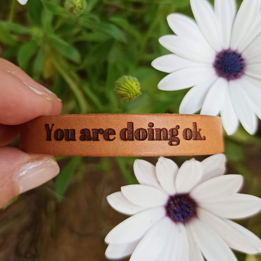A brown leather bracelet with the text 'You are doing ok.' engraved on it.