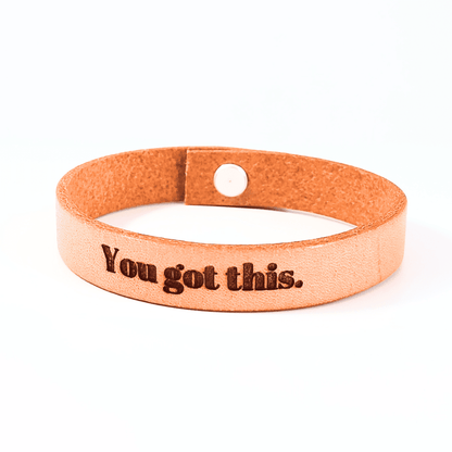 A brown leather bracelet with the text 'You got this.' engraved on it.