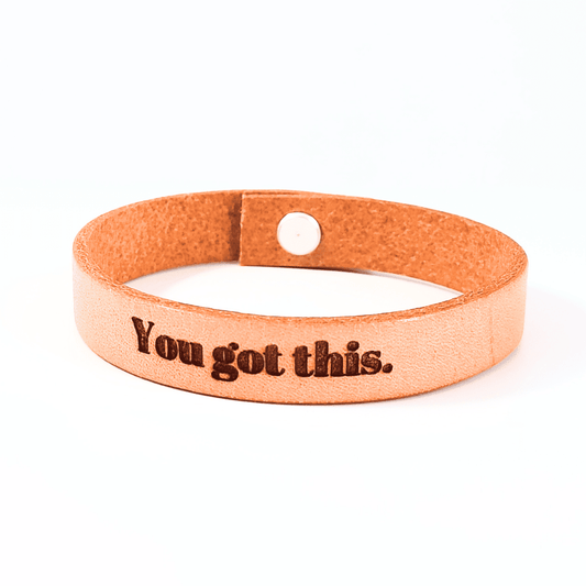 A brown leather bracelet with the text 'You got this.' engraved on it.